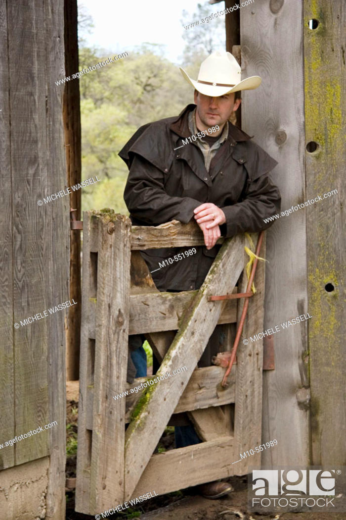 Stock Photo: Cowboy up against gate and old rugged barn in the country on a ranch looking downward in a relaxed style.