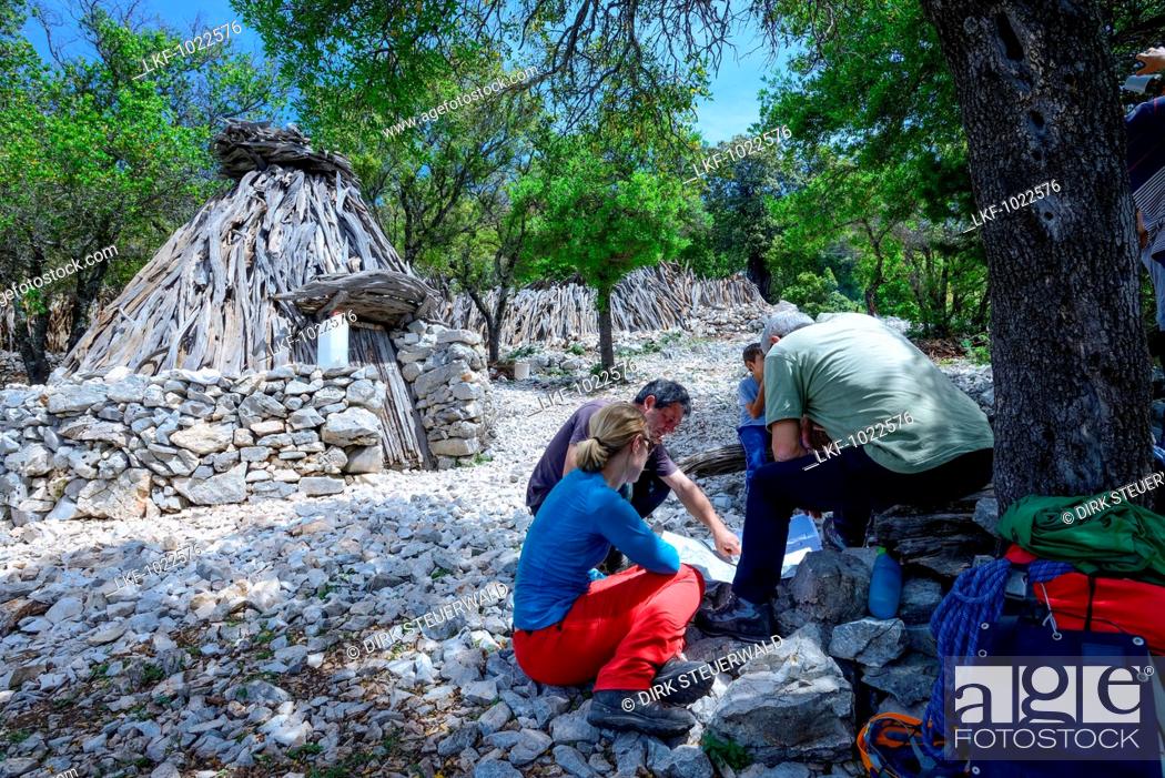 Stock Photo: A young woman with trekking gear discussing the trail on a map with locals in front of an Ovile, which is a traditional shepherds hut, Selvaggio Blu, Sardinia.