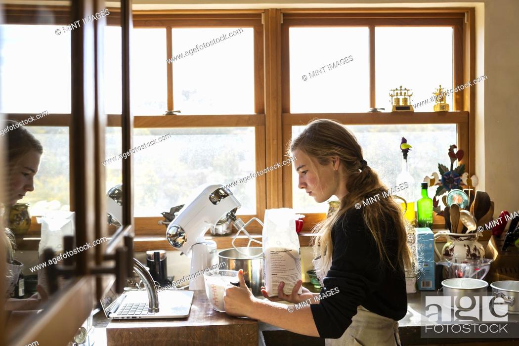 Photo de stock: Teenage girl in a kitchen following a baking recipe on a laptop.