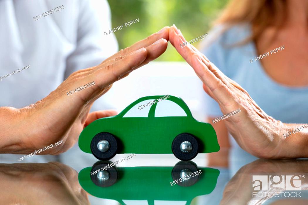 Stock Photo: Close-up Of Couple's Hand Protecting Green Wooden Car Over Reflective Desk.