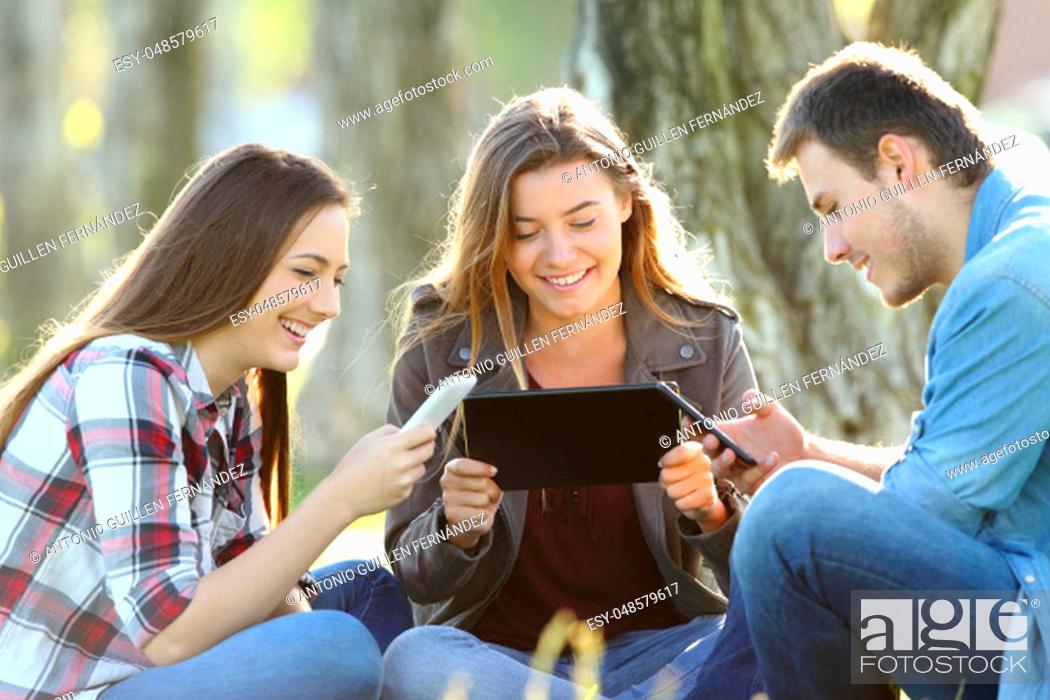 Stock Photo: Three happy friends using multiple devices sitting on the grass in a park.