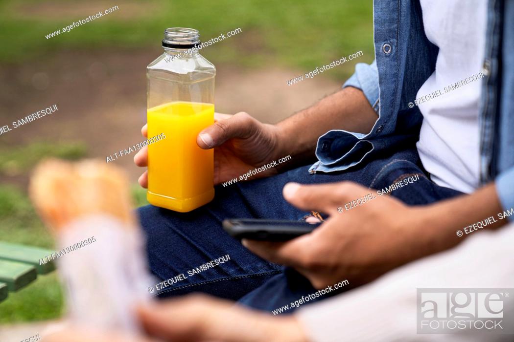 Photo de stock: Mid-shot of man's hand holding an orange juice bottle and an out-of-focus hand holding a sandwich.