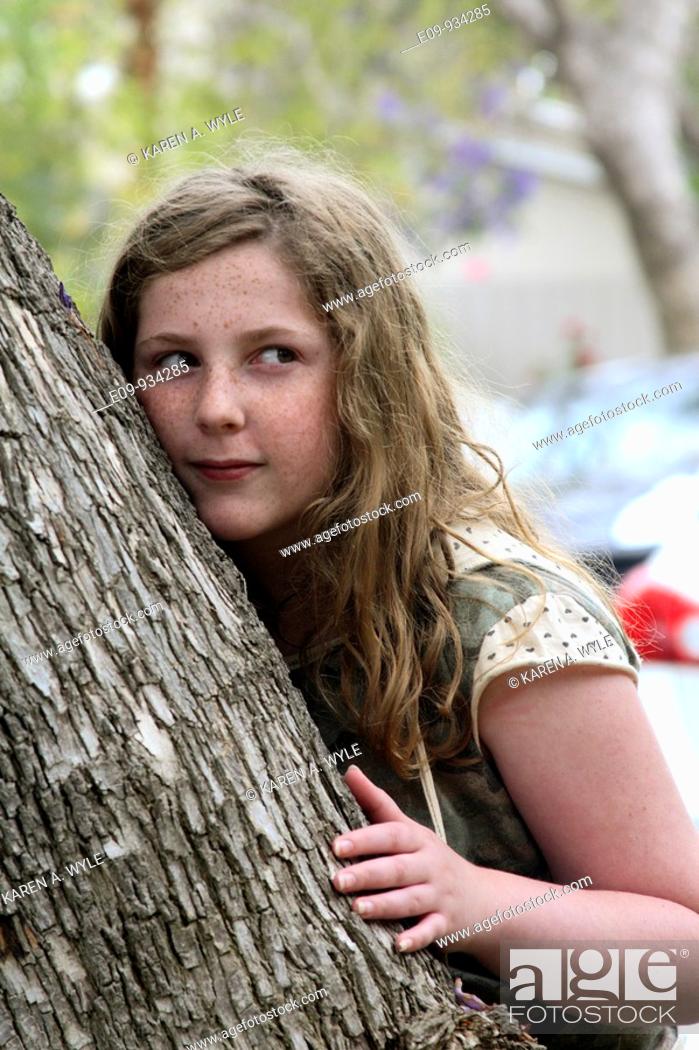 Stock Photo: 13-year-old girl with blonde-brown long hair and freckles, half leaning against, half hiding behind a tree, up-to-something expression.