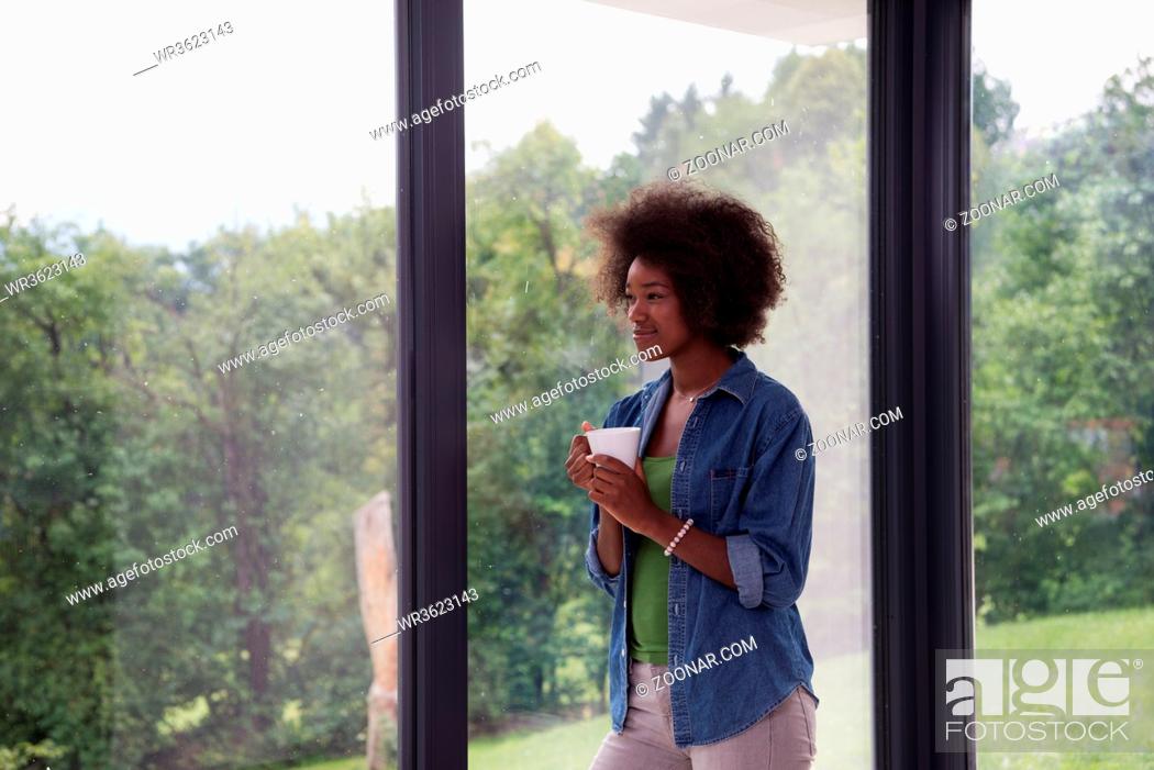 Stock Photo: Beautiful young african american woman drinking coffee and looking through a window in her luxury home.