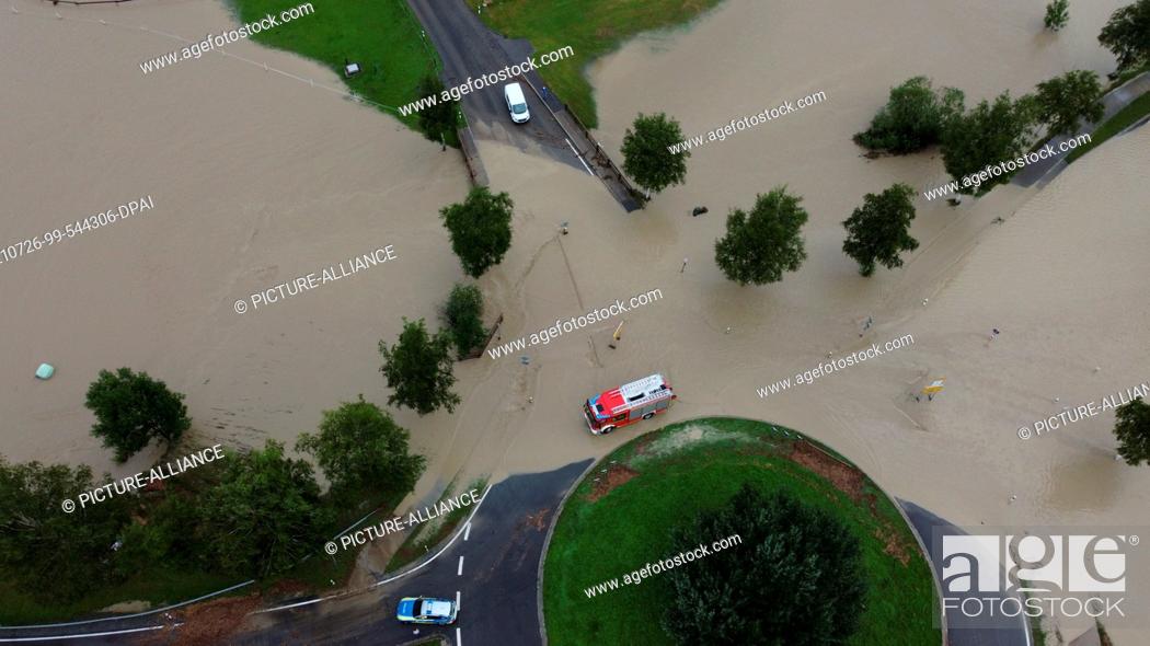 Stock Photo: 26 July 2021, Bavaria, Rettenberg: A fire engine drives through the high standing water at a roundabout outside the town (shot with a drone).