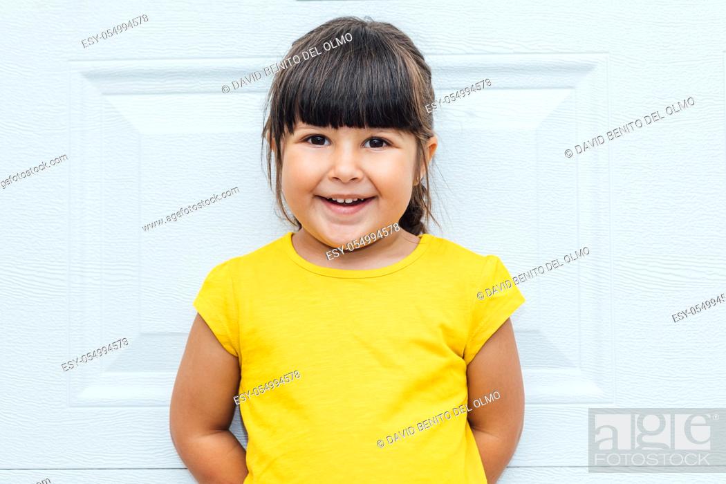 Stock Photo: Adorable little girl with black hair wearing a yellow shirt leaning against white background.