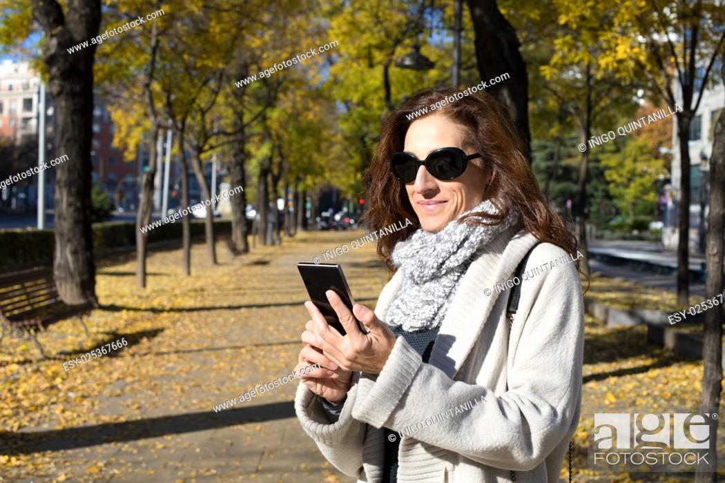 Photo de stock: happy woman with grey cardigan and scarf, black sunglasses, watching smartphone in urban street in Madrid city with trees in autumn.