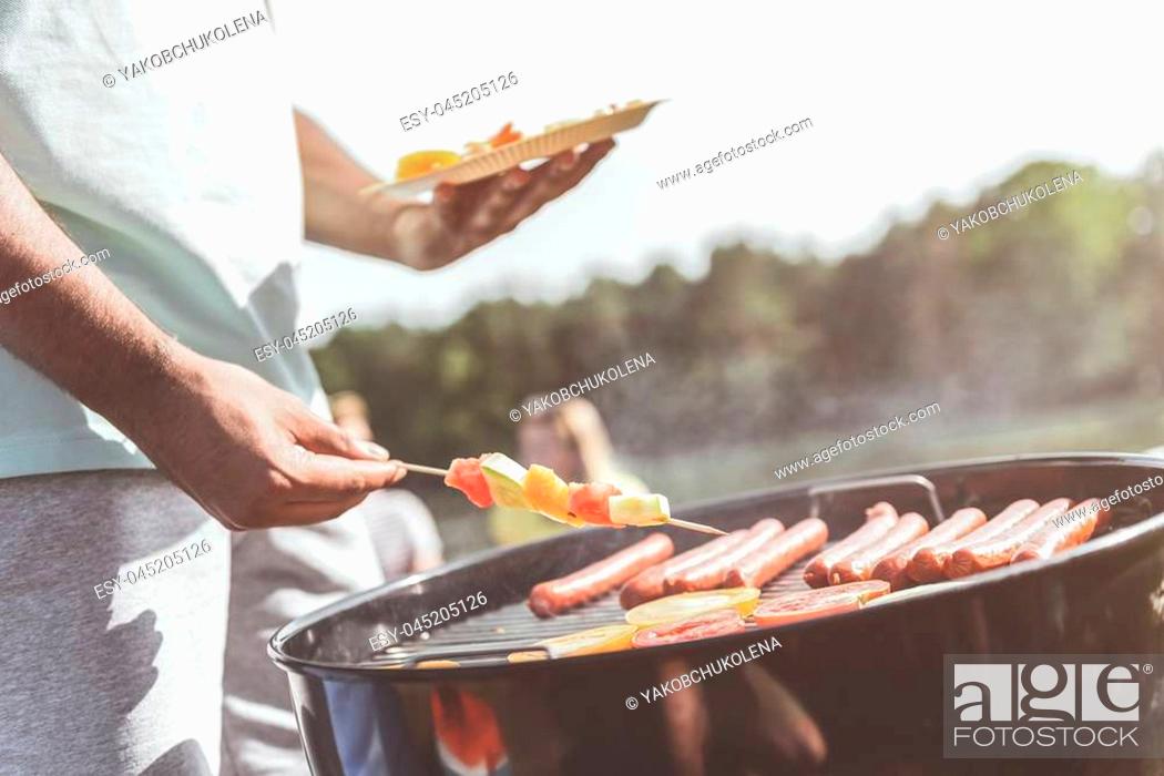 Stock Photo: male hands holding vegetable skewers and plate while standing near grill. Vegies and sausages are on grate. Close up.