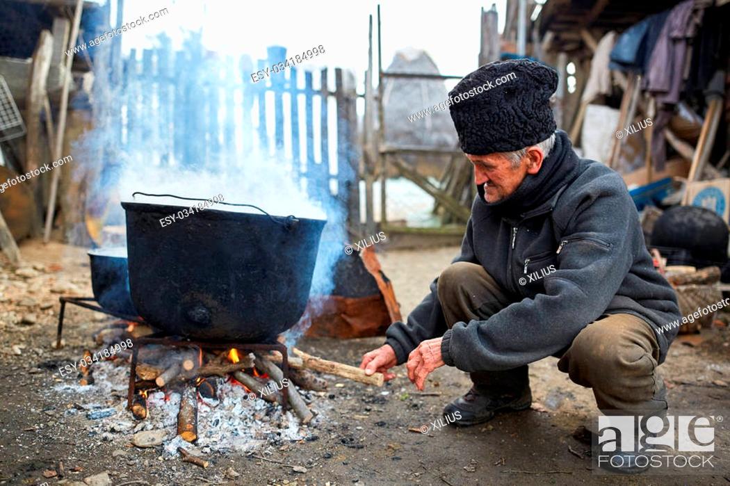 Stock Photo: Old man putting wood on fire below large boiling cast iron pots.