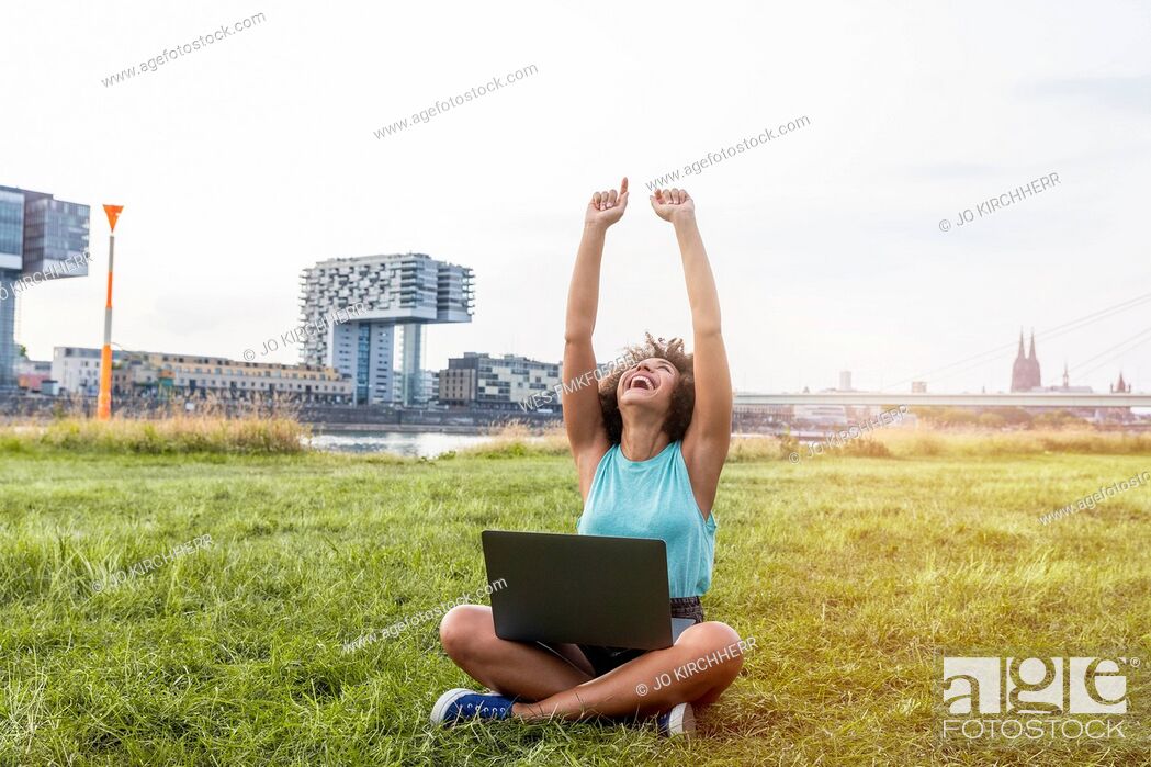 Stock Photo: Germany, Cologne, cheering woman sitting on a meadow using laptop.