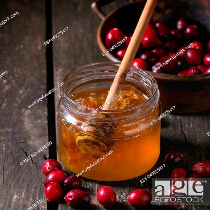 Stock Photo: Open glass jar of liquid honey with honeycomb and honey dipper inside and fresh cranberries in vintage bowl over old wooden table. Dark rustic style.