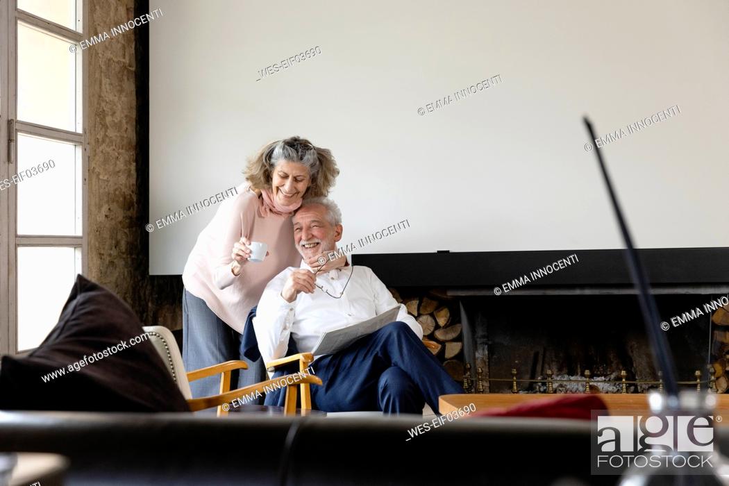 Stock Photo: Happy senior woman holding coffee cup embracing man sitting with newspaper in living room at home.