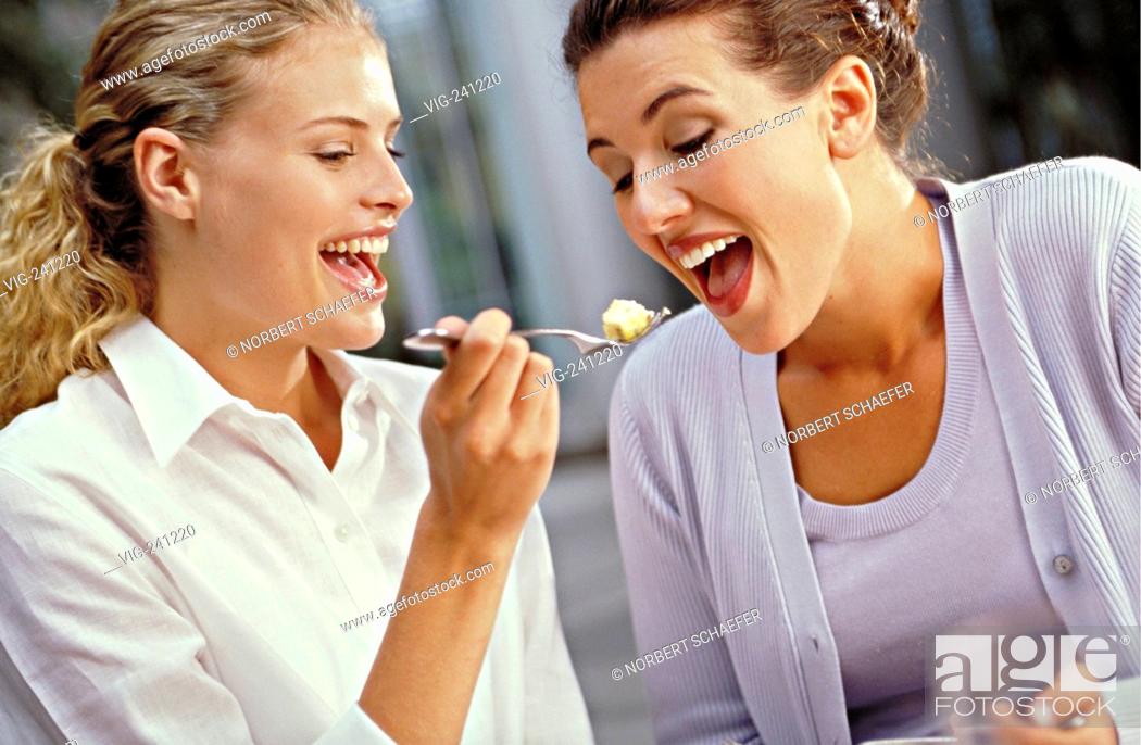 Stock Photo: portrait, young blond curly woman balances in a bar a piece of cheese on a fork into the mouth of her brunette girl friend  - GERMANY, 27/01/2004.