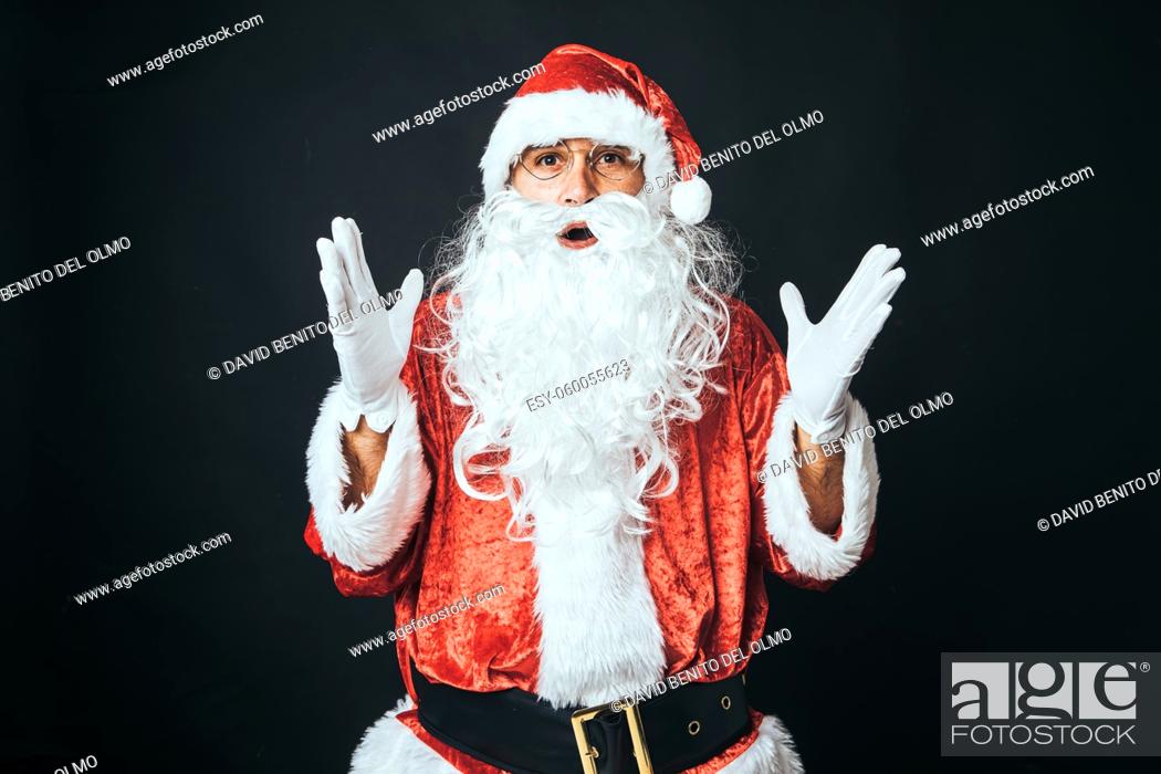Stock Photo: Man dressed as Santa Claus surprised, with raised hands, on black background. Christmas concept, Santa Claus, gifts, celebration.