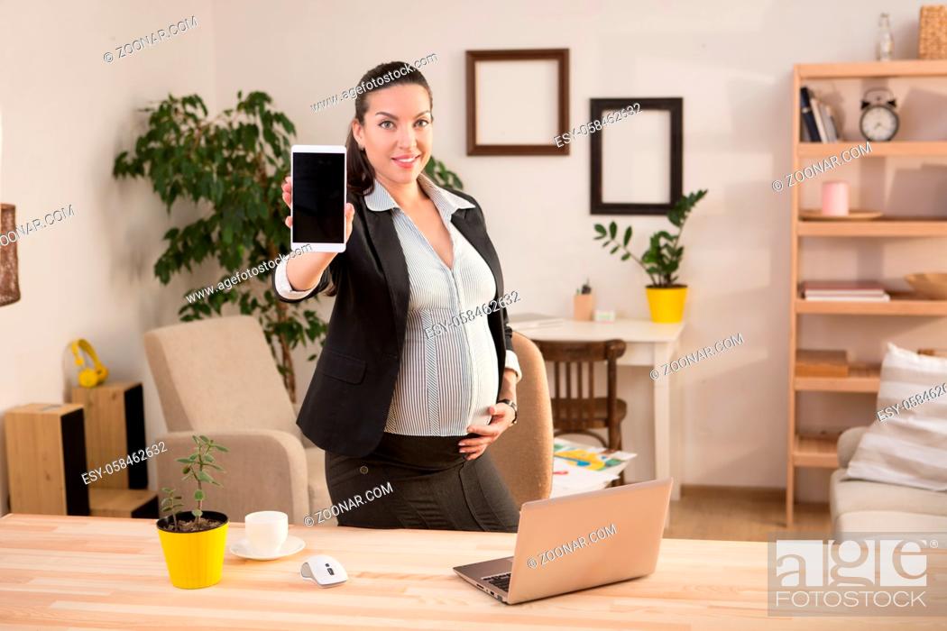 Stock Photo: Portrait of happy and cheerful pregnant business woman smiling and demonstrating her mobile or smart phone to camera while working at home.