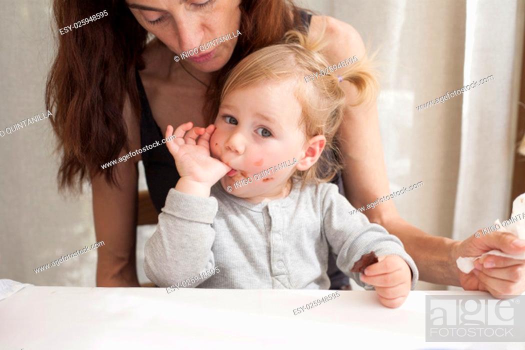 Stock Photo: portrait of two years age blonde baby with grey shirt sucking her thumb finger eating chocolate piece sitting on legs of mother woman black shirt in white table.
