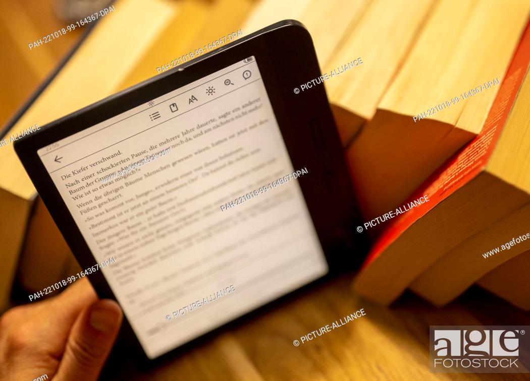 Stock Photo: ILLUSTRATION - 17 October 2022, Berlin: A man holds an e-book reader in his hand while numerous paper books lie on the table in the background.