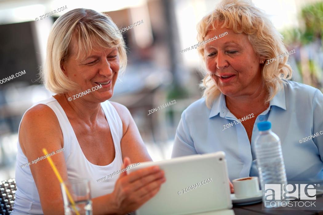 Stock Photo: Two smiling senior women are sitting close to each other at a table of an outdoor cafe. They are both looking at the tablet that one of the women is holding in.