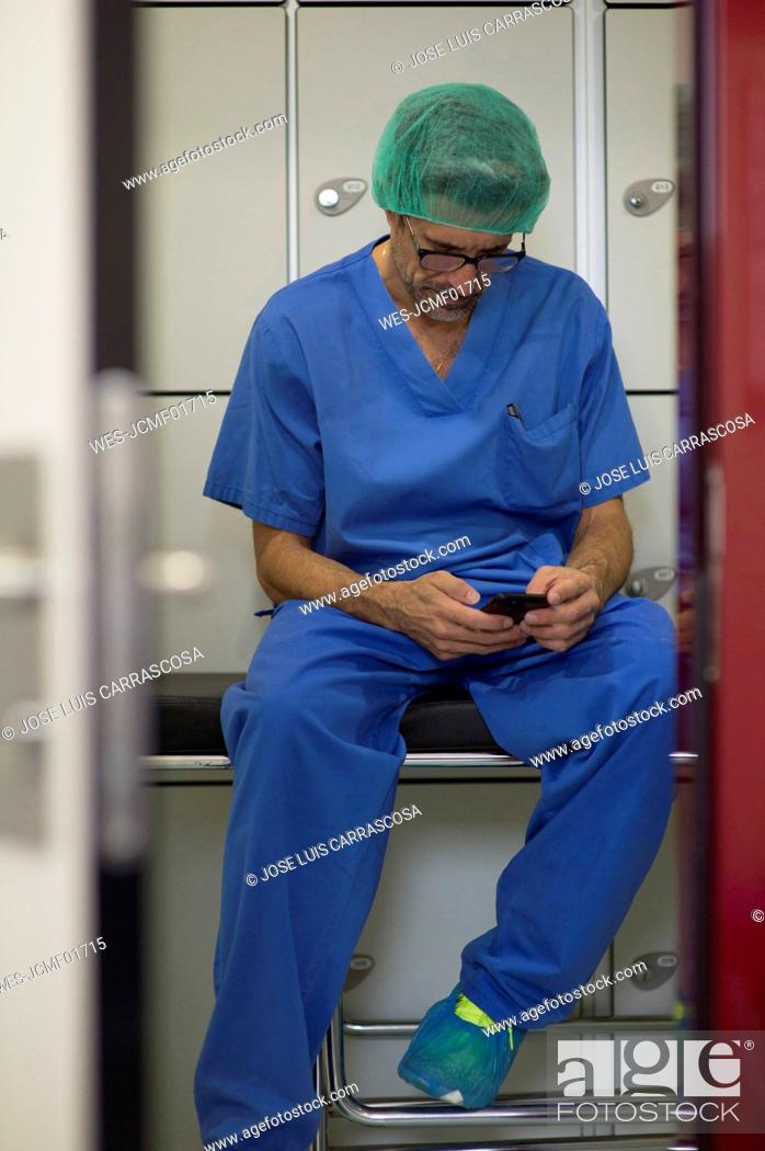 Stock Photo: Male surgeon in surgical cap using smart phone at hospital.