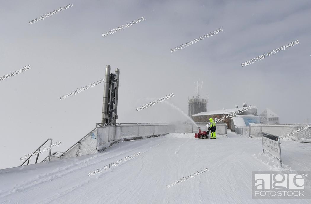 Stock Photo: 07 October 2021, Bavaria, Grainau: An employee clears the roof terrace of the Zugspitze mountain station with a snow blower.