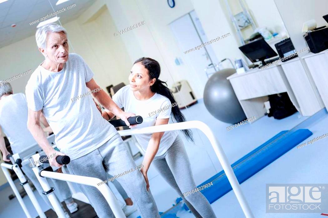 Stock Photo: Much better. Positive emotional aged man feeling confident while being in a medical center and trying to make a few steps on a convenient treadmill with a help.