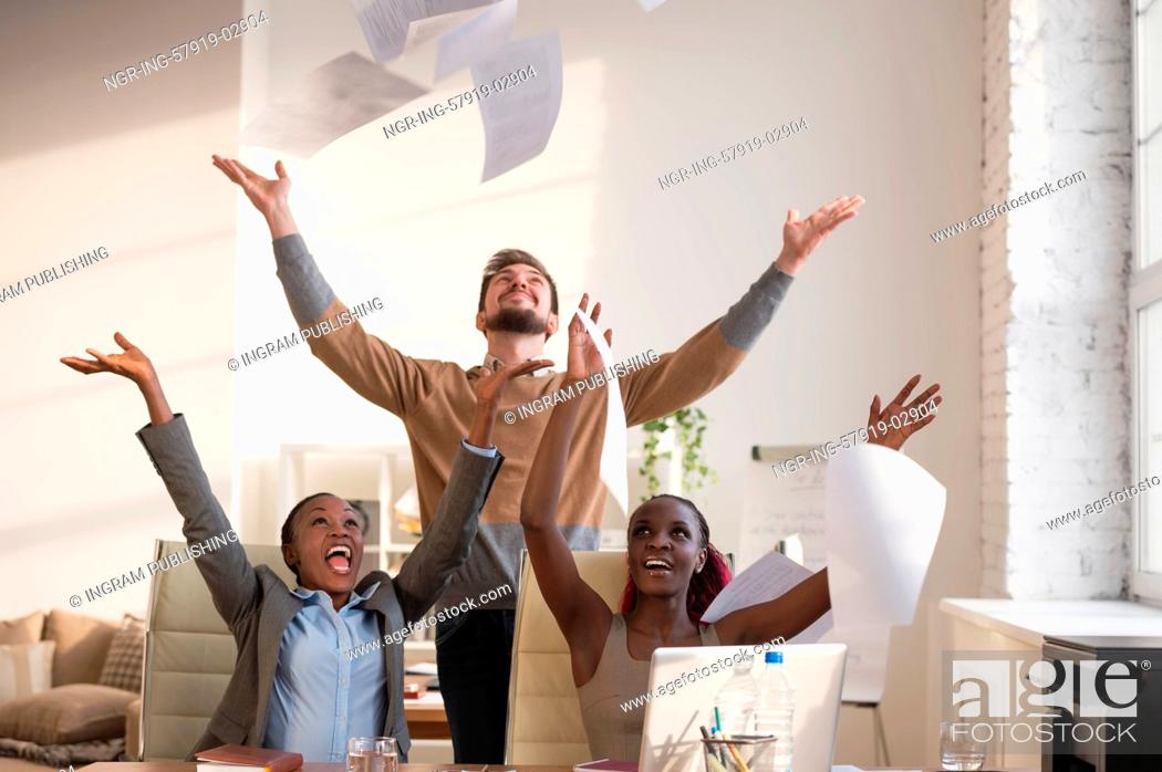 Stock Photo: Business people excited happy smile, throwing up papers, documents fly in air, businesspeople sitting at office desk hold hands arms up, success team concept.