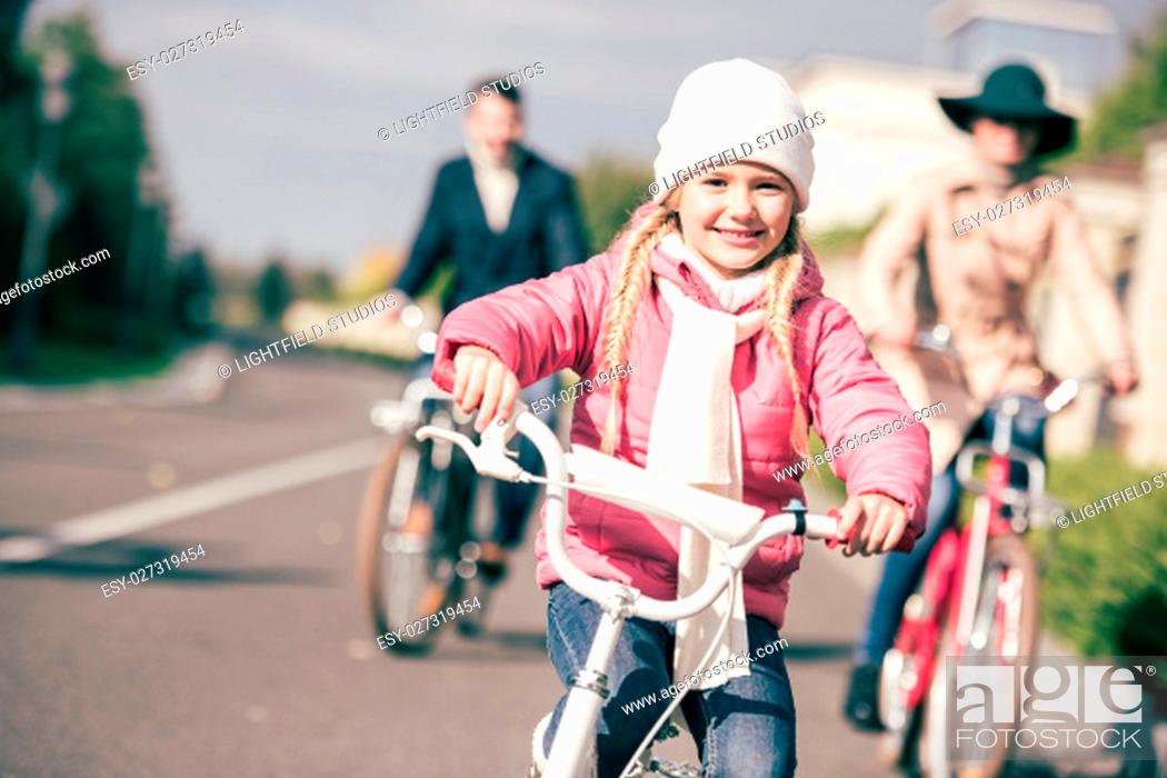 Stock Photo: Close-up portrait of cute little girl riding bicycle with blurred parents on background.