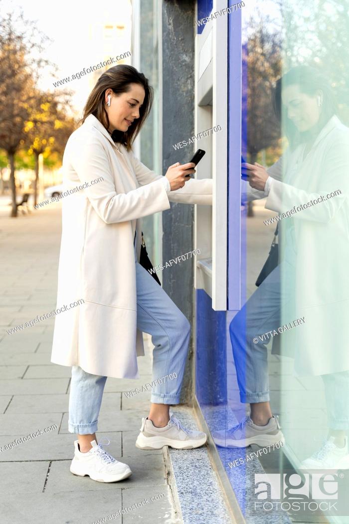 Stock Photo: Stylish female entrepreneur using smart phone while withdrawing money from ATM.