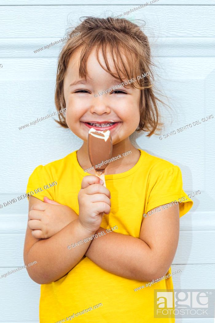Stock Photo: Adorable blond-haired girl eating ice cream, wearing a yellow shirt leaning against white background.