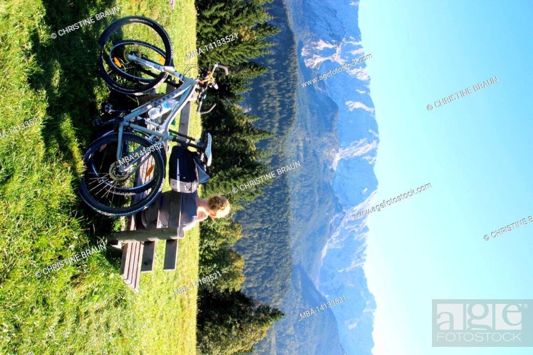Stock Photo: young woman takes a break on a bicycle tour on a bench at the eckbauer in front of the wetterstein massif with a wonderful blue sky, autumn in werdenfelser land.