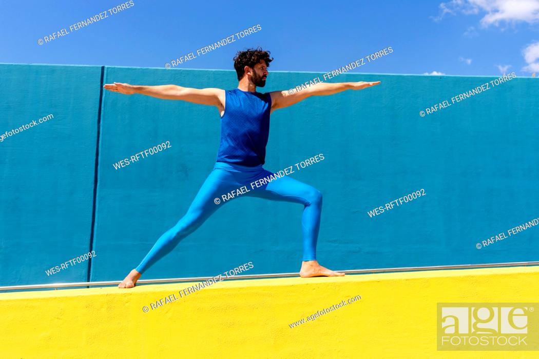 Stock Photo: Mid adult man practicing Virabhadrasana II on retaining wall during sunny day.