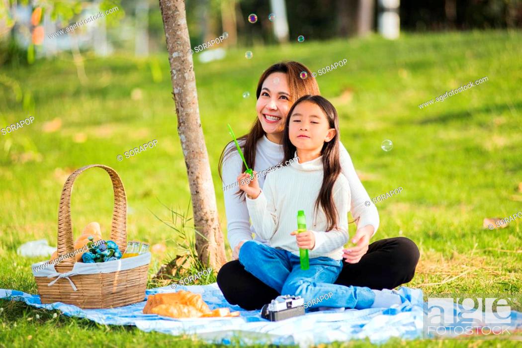Photo de stock: Happy Asian mother and little girl daughter child having fun and enjoying outdoor together sitting on the grass blowing soap bubbles during a picnic in the.