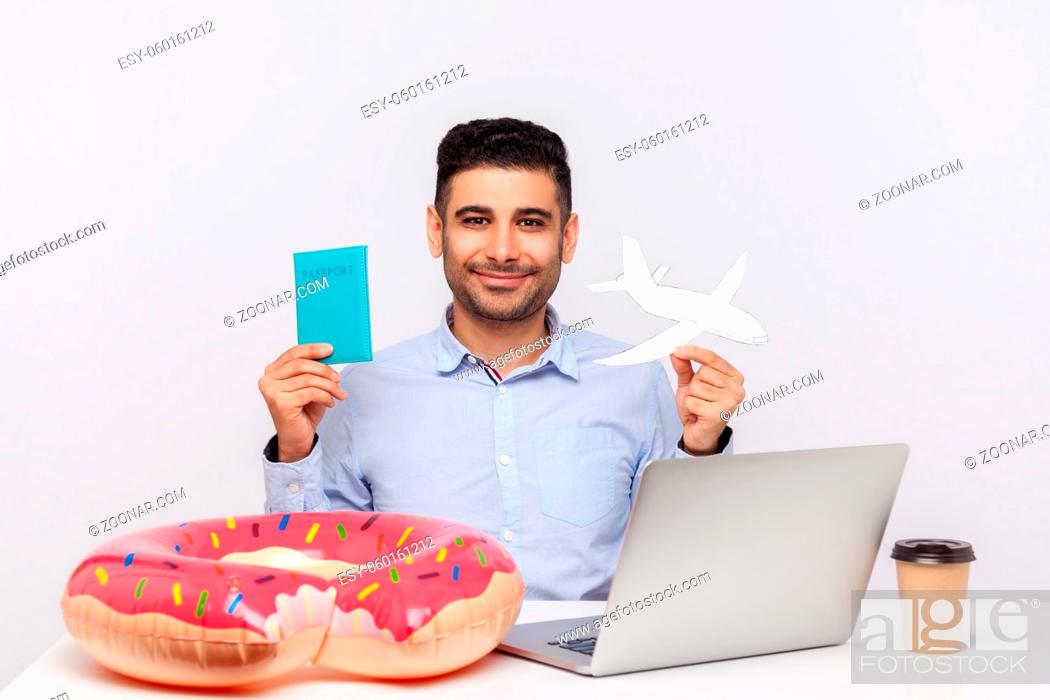 Photo de stock: Cheerful man holding paper airplane and passport in hands, sitting in office workplace with rubber ring on desk, planning travel vacation, weekend trip.
