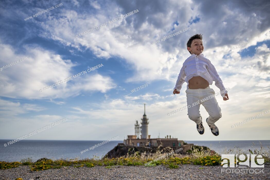 Photo de stock: Child jumps for photo one day with clouds with lighthouse and sea in background.