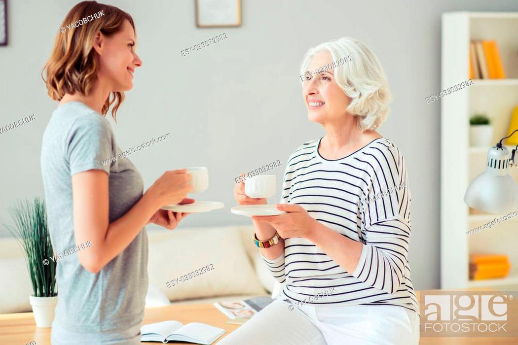 Stock Photo: Get some energy. Positive delighted woman leaning on the table and drinking tea while resting together with her adult charming daughter.