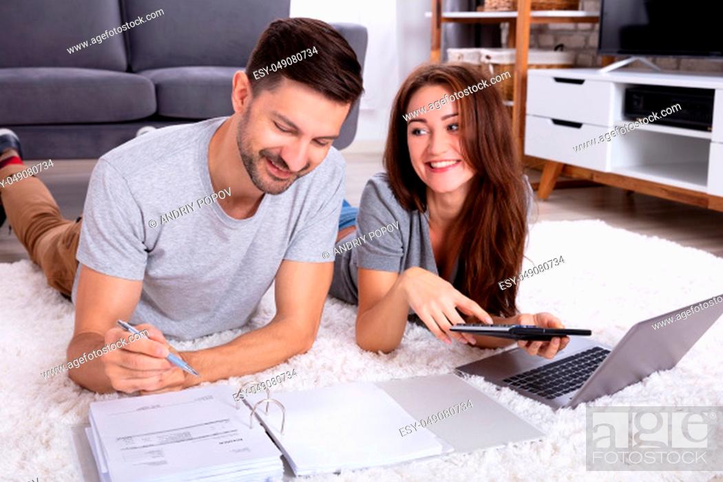 Imagen: Smiling Young Couple Lying On Carpet Calculating Invoice With Calculator.