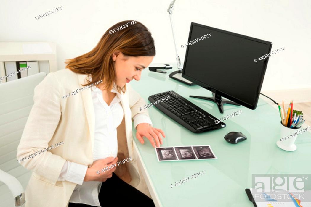 Stock Photo: Happy Pregnant Businesswoman Looking At Ultrasound Scan Report On Desk.