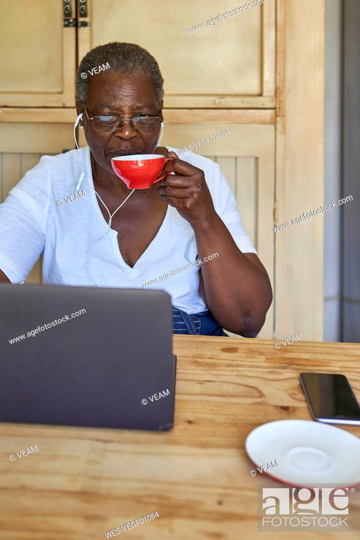 Stock Photo: Senior woman sitting at kitchen table using laptop and drinking from cup.
