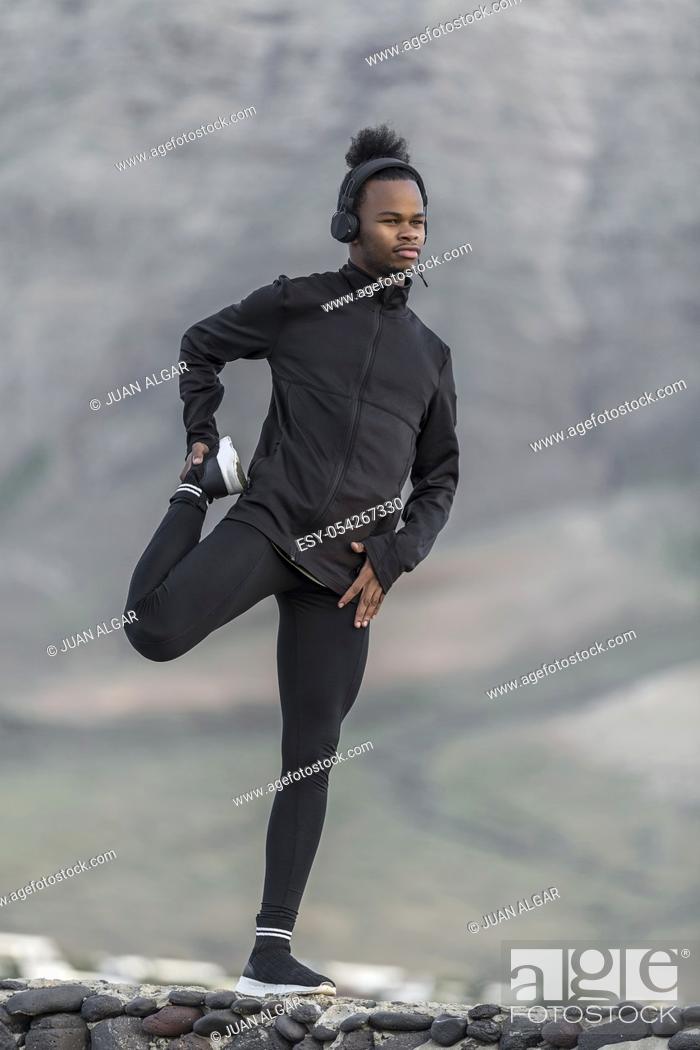Stock Photo: Handsome African American male in sportswear listening to music and doing warm up exercise for legs while standing on stones on blurred background of nature.