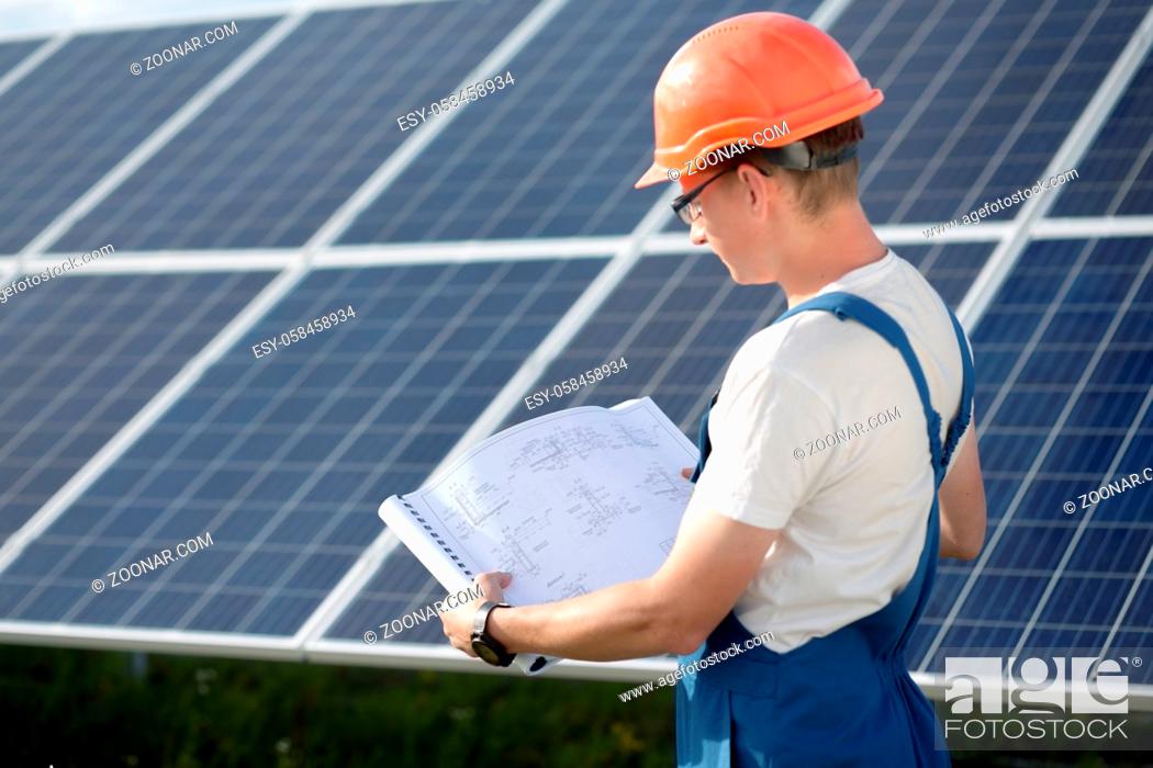 Stock Photo: Young worker in overall looking in drawings at solar power station. Employee in blue overall and orange helmet, looking in technical documentation.