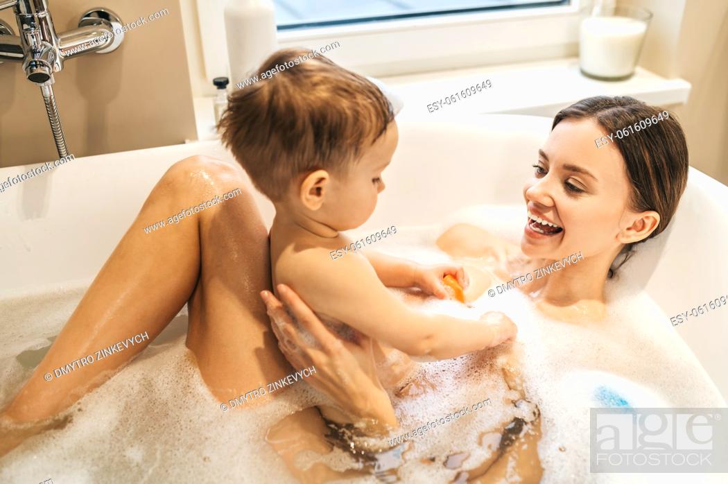 Stock Photo: Joyous cute young female parent lying in the bathtub and playing with her baby boy.