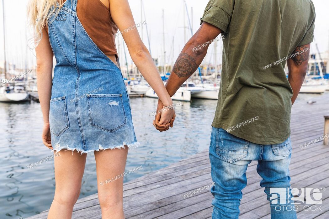 Imagen: Back view of multicultural young couple standing hand in hand on jetty, partial view.