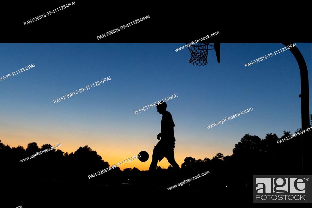 Stock Photo: 16 August 2022, Bavaria, Nuremberg: A youth plays basketball at a public hoop under a bridge at dusk. Photo: Daniel Karmann/dpa.