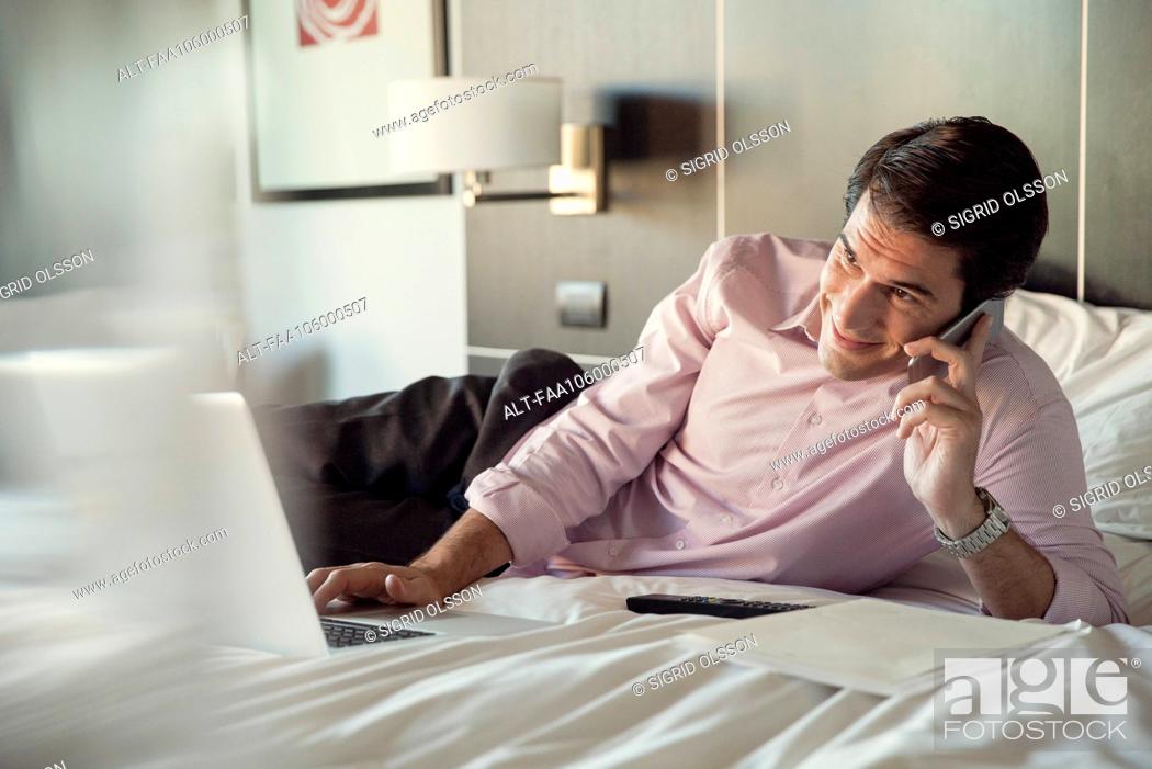 Stock Photo: Man lying on hotel bed, talking on cell phone and using laptop computer.
