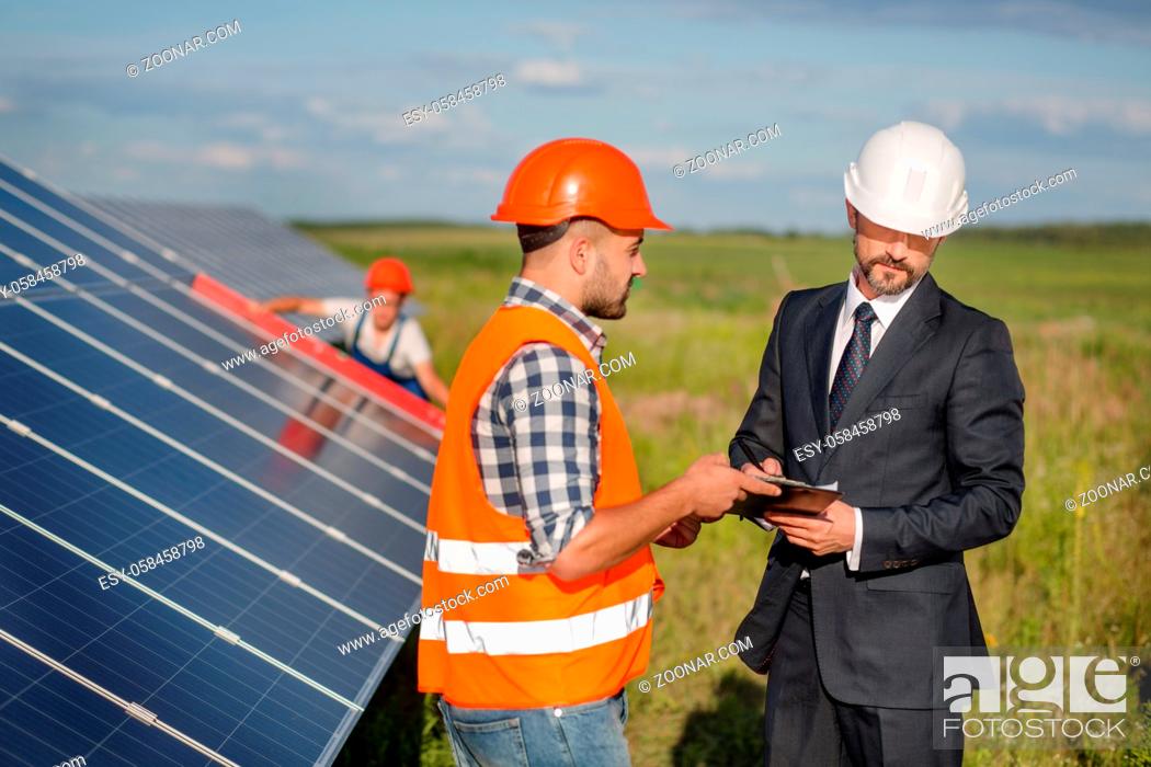 Stock Photo: Businessman signing contract with foreman, technician examining solar panels at backdrop. Signing of agreement at solar energy station in the field.