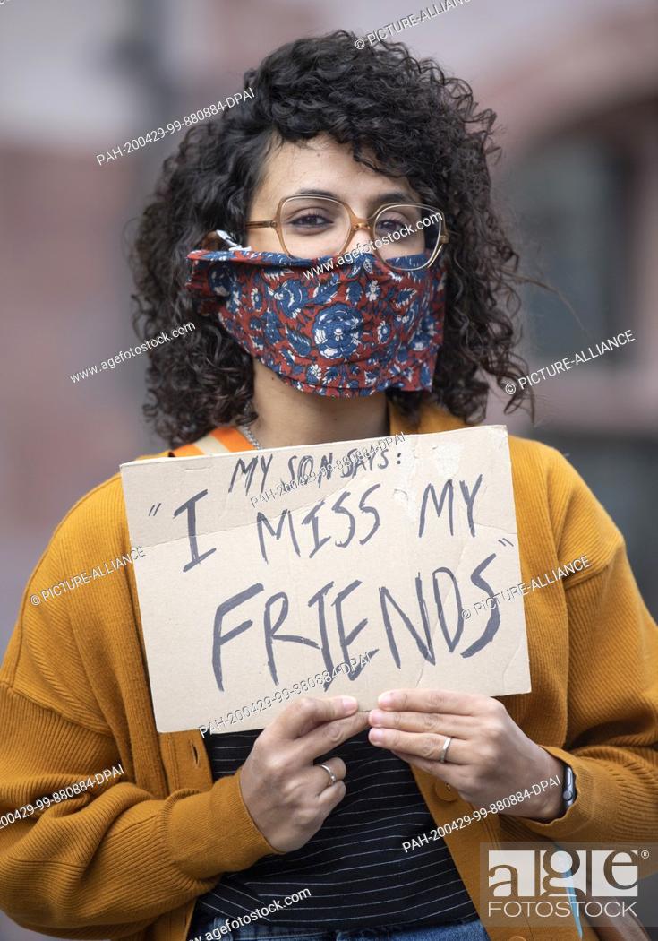Stock Photo: 29 April 2020, Hessen, Frankfurt/Main: A young woman who has her nose and mouth covered shows a poster with the inscription ""My son says: I miss my friends"".