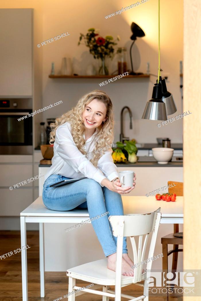 Stock Photo: Smiling blond freelancer holding coffee cup sitting with laptop on table in kitchen at home.