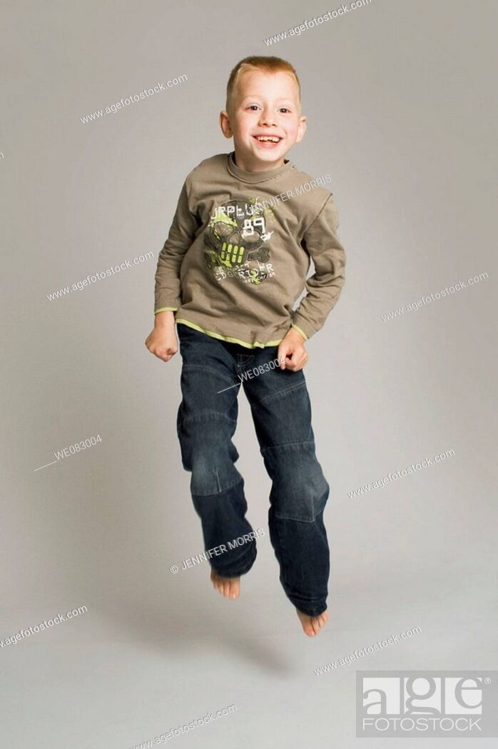 Stock Photo: Blonde-haired, blue-eyed six year old boy in studio, jumping and smiling against a pale grey background. He wears jeans and a long-sleeved t-shirt.