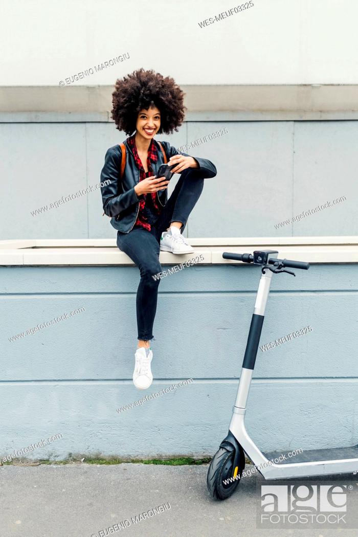 Imagen: Smiling woman holding mobile phone while sitting on retaining wall by electric push scooter.