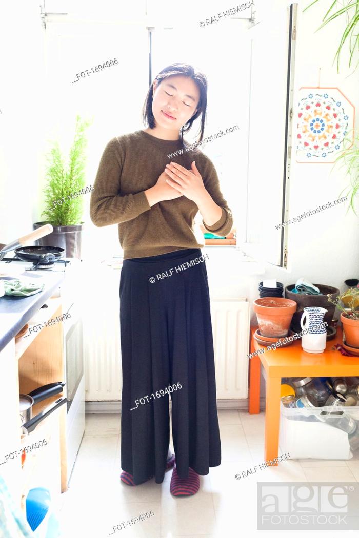 Photo de stock: Full length of young woman with hands clasped standing against window in kitchen.